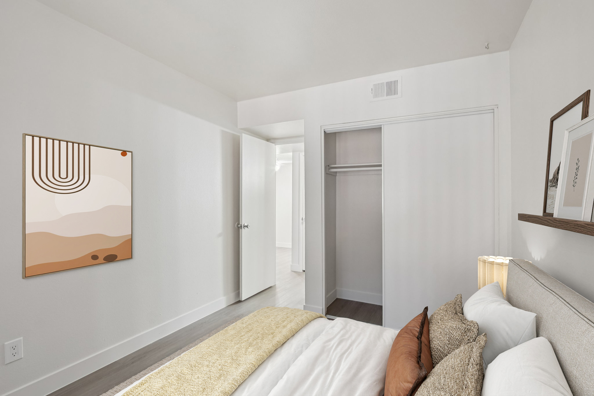 A cozy bedroom featuring a neatly made bed with decorative pillows, a light-colored comforter, and a textured throw. The wall displays an abstract art piece in earthy tones. An open closet is visible, alongside a doorway leading to another room, with soft lighting creating a warm atmosphere.