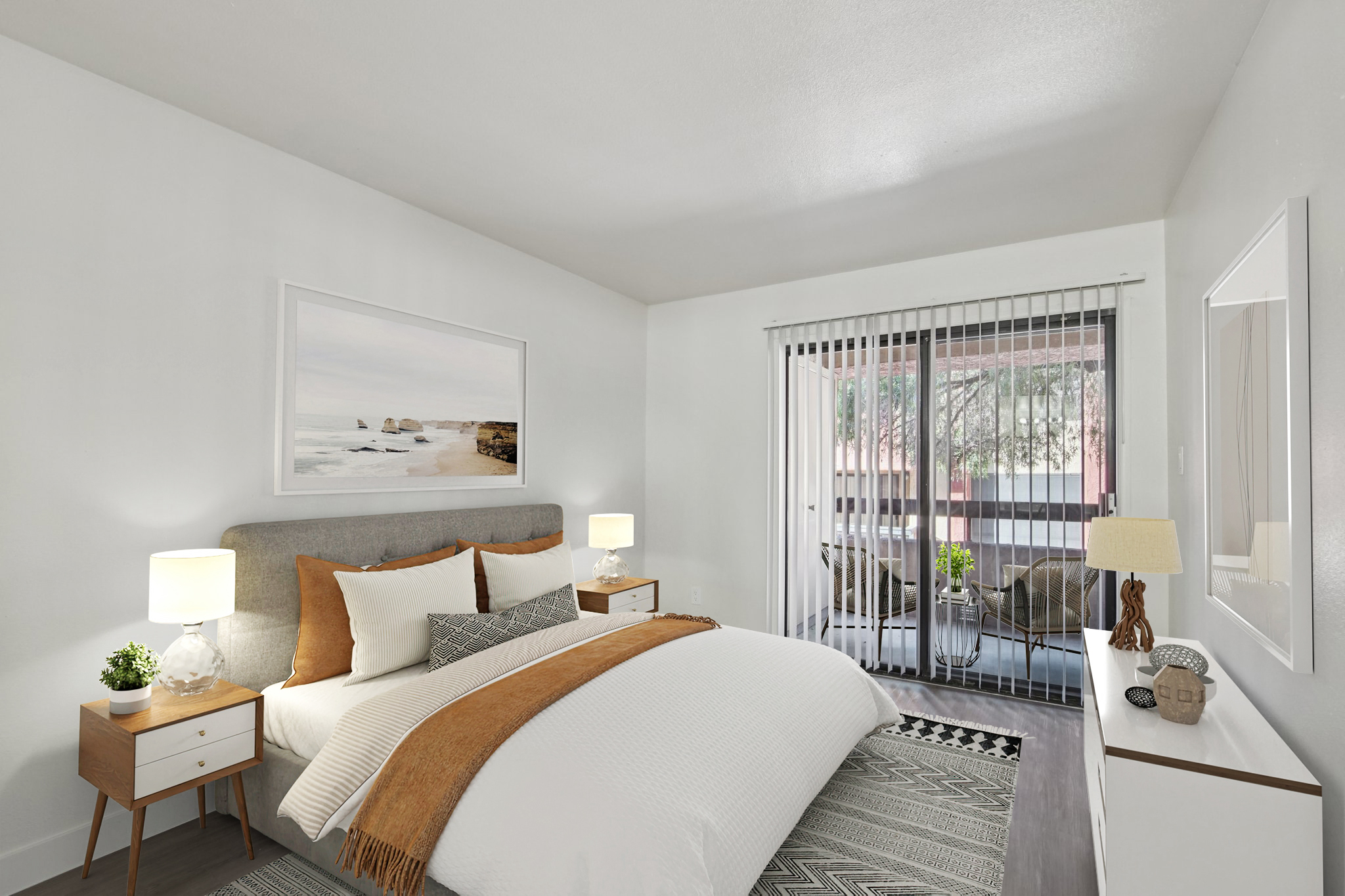 A modern bedroom featuring a queen-sized bed with a gray upholstered headboard and decorative pillows. There are two bedside tables with lamps, a large framed landscape photo on the wall, and sliding glass doors leading to a small balcony with outdoor seating. The room has a clean, minimalist aesthetic.