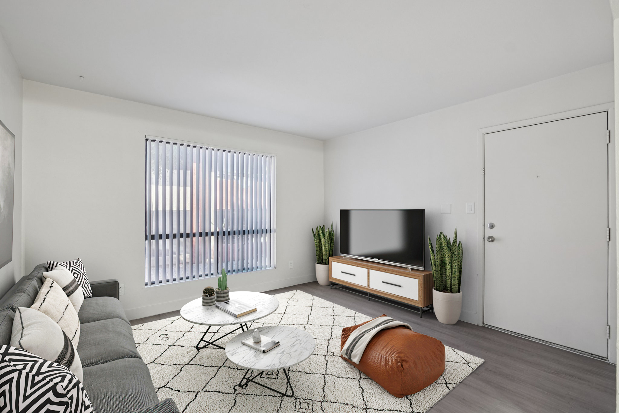 A modern living room featuring a gray sofa with decorative pillows, a round coffee table with a marble top, and a large flat-screen TV on a wooden console. There are potted plants in the corner, a large window with vertical blinds, and a cozy, patterned area rug on the floor. The walls are painted in a light color.