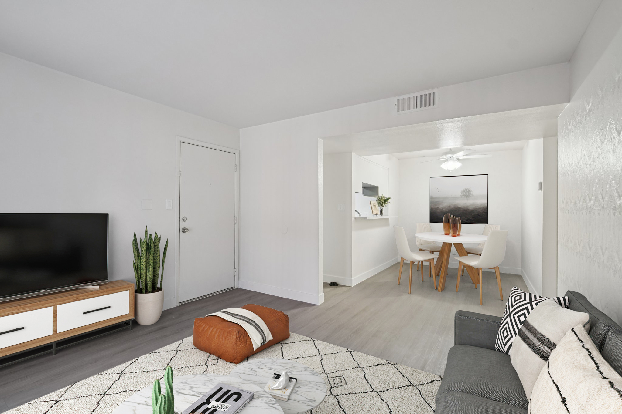 A bright, modern living room featuring a gray sofa, a large television on a wooden media console, and a geometric-patterned rug. A stylish dining area with a round table and white chairs is visible in the background, along with a potted plant and decorative accessories. The walls are painted white, creating an open and airy atmosphere.