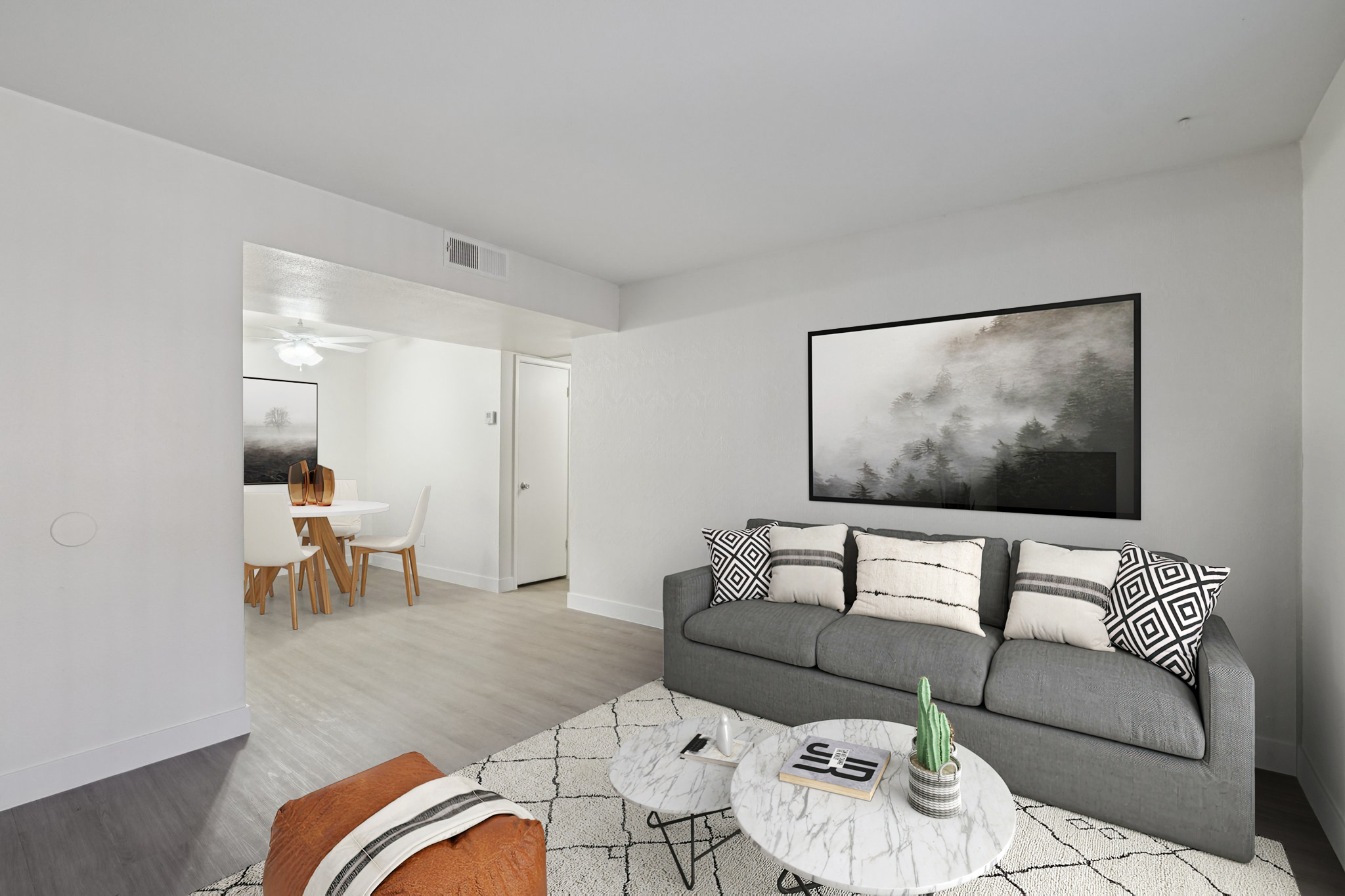 A modern living room featuring a gray sofa with striped and patterned cushions, a round coffee table with a plant, and a textured rug. A large wall art piece is displayed above the sofa. In the background, a dining area with a round table and chairs is visible, illuminated by natural light.