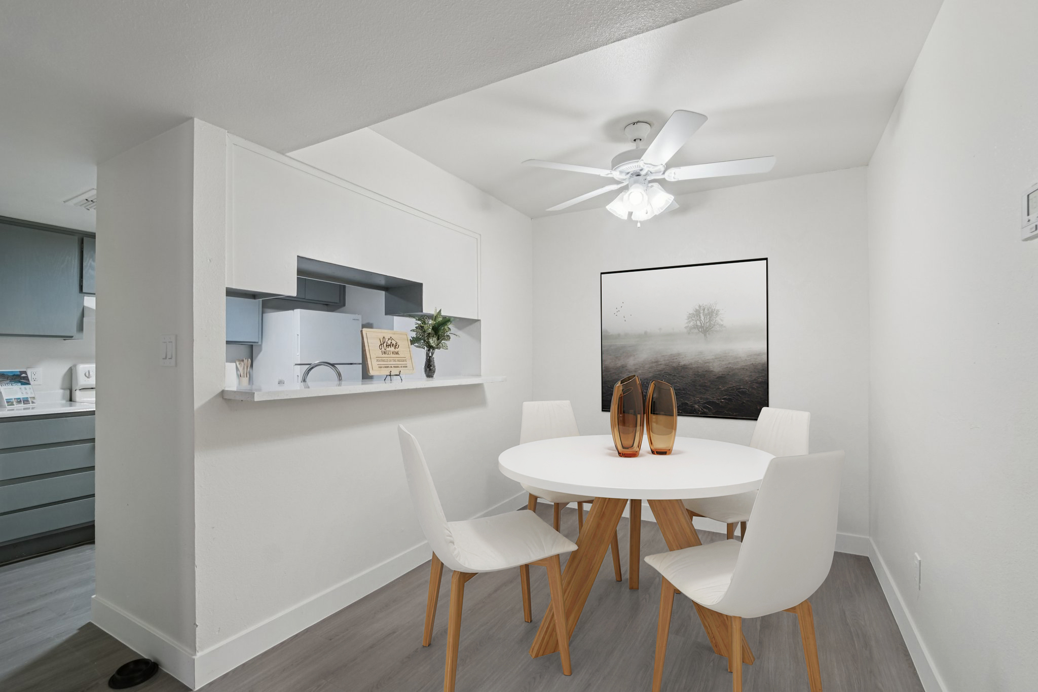 A modern dining area featuring a round white table surrounded by four white chairs. In the background, there's a small kitchen area with grey cabinets. A framed artwork of a misty landscape is on the wall, and a ceiling fan illuminates the space. The flooring is light-colored, enhancing the bright atmosphere.