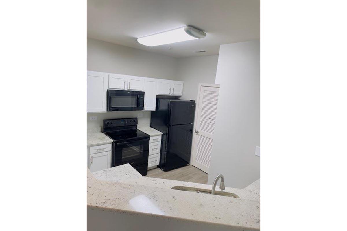 A modern kitchen featuring white cabinets, black appliances including a microwave, stove, and refrigerator, and a granite countertop. The area has a bright overhead light and a view from a slightly elevated angle, showcasing an open design with an adjoining space.