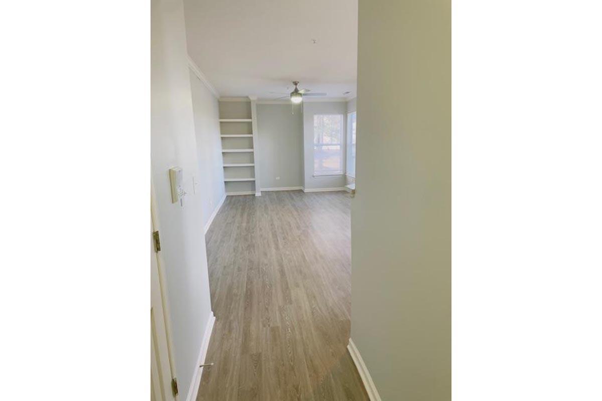 A spacious, light-filled room with light wooden flooring and a ceiling fan. To the left, there is an open doorway leading into another area, and to the right, a large window offers natural light. Shelves are visible along the wall in the background, suggesting storage or display space.