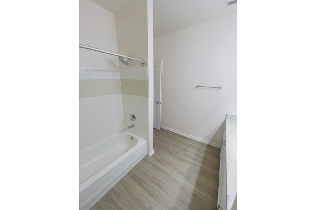 A bathroom featuring a white tub with a shower attachment, light-colored walls, and a countertop with a single sink. The flooring is light wood, and there is a towel bar on the wall. A door leads to another area, creating a spacious and clean aesthetic.