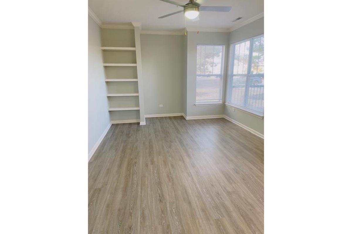 A light-filled, empty room with a ceiling fan, featuring wooden flooring and large windows. One wall has a built-in shelf unit. The walls are painted in a soft, neutral color, creating a spacious and inviting atmosphere.