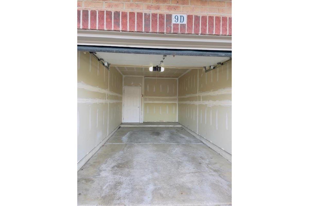Empty garage with beige walls and a concrete floor. A single white door is visible at the back, and the garage door is partially open. The overhead lighting is off, and there are no vehicles or items present in the space. The address number "9D" is displayed above the garage entrance.