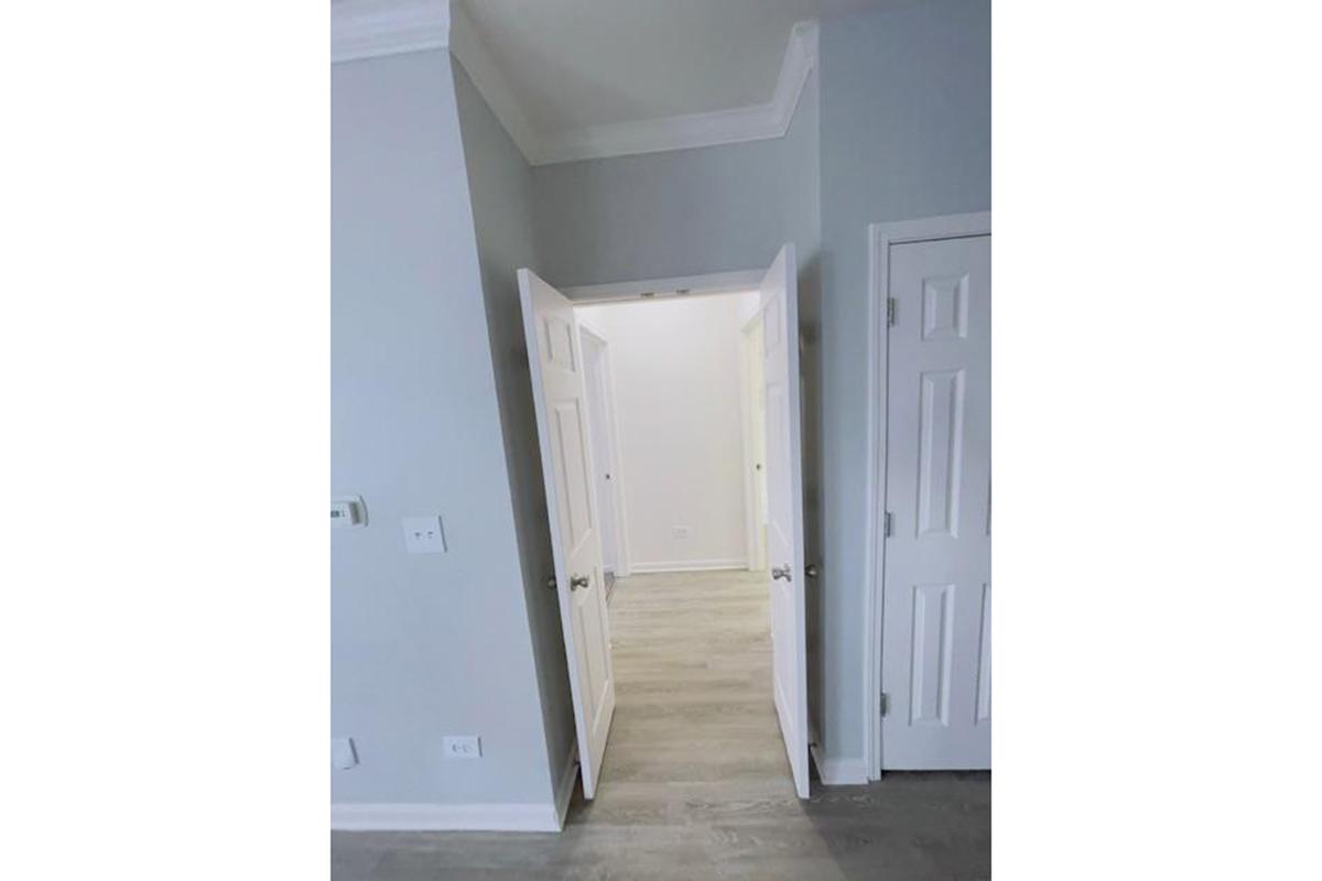 A well-lit interior space featuring light gray walls and a light-colored floor. Two doors are visible, one leading to a room and the other likely to a closet. The area appears clean and spacious, with a simple design and a ceiling with crown molding.