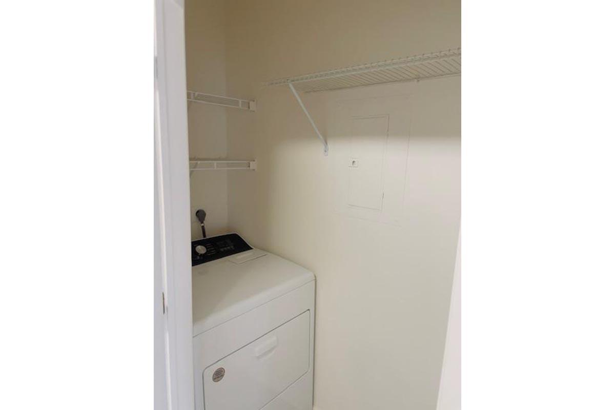 Interior view of a small laundry room featuring a white washing machine and a shelf for storage. The walls are painted a light color, and there is a utility panel visible. The space has minimal decor, providing a clean and functional utility area.