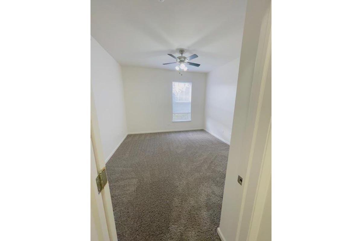 A vacant room with light gray carpet and white walls. The room features a ceiling fan with three lights and a window allowing natural light to enter. The space appears clean and ready for furniture, offering a neutral backdrop for various decorating styles.