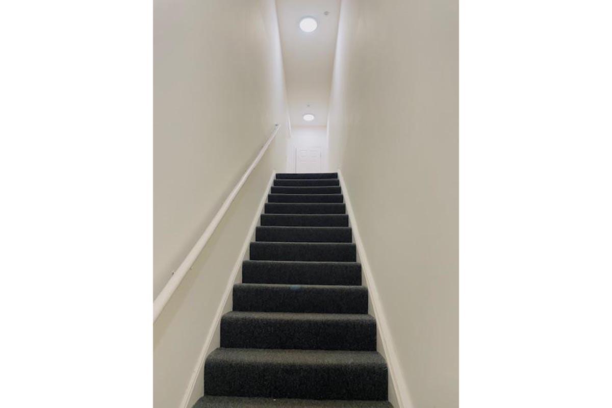 A narrow staircase with dark carpet leading upwards, featuring a plain white wall on either side. Soft lighting from circular ceiling fixtures illuminates the space, and a closed door is visible at the top of the stairs.