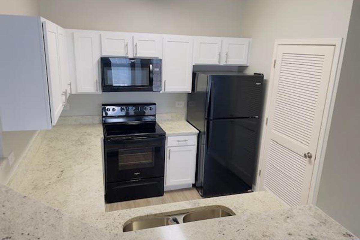 A modern kitchen featuring white cabinets, a black refrigerator, an oven, and a microwave. The countertops are light colored, with a double sink visible. The layout is open and spacious, with a neutral color scheme and ample natural light.