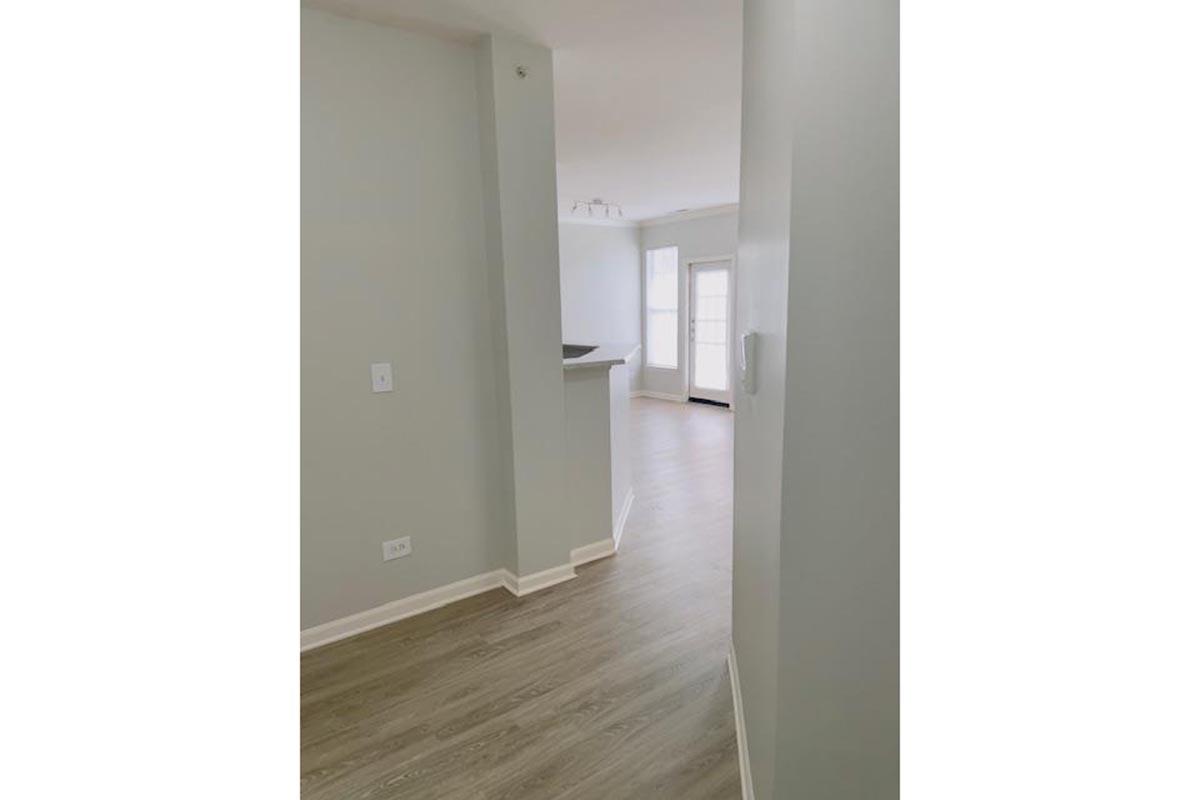 A well-lit hallway leading to an open living area, featuring light gray walls and a light wood floor. The space has a modern design with natural light streaming in from a large window at the end, and a white wall on the left with a small partial wall near the entryway.