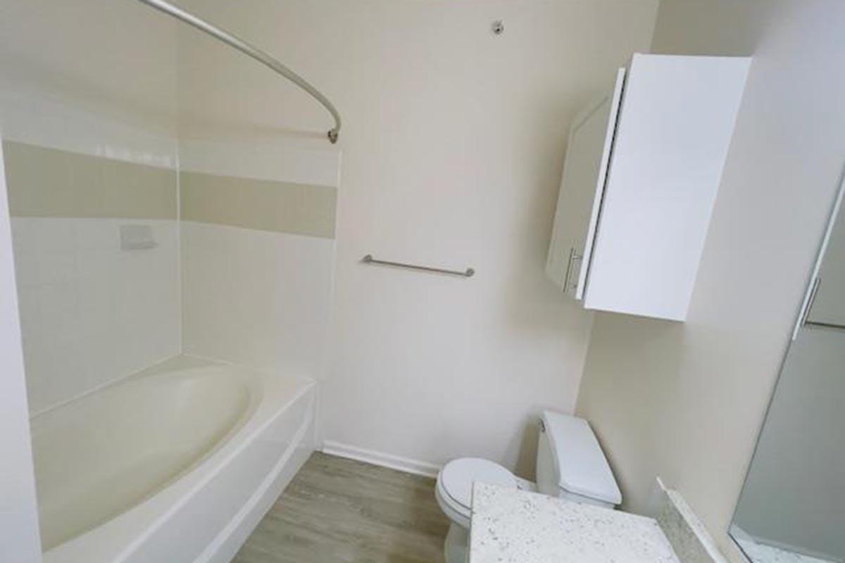 A bright, clean bathroom featuring a tub with a shower curtain rod, a toilet, and a small vanity with a white cabinet. The walls are light-colored, and the flooring is a neutral tone. A large mirror is above the sink, enhancing the spacious feel of the room.