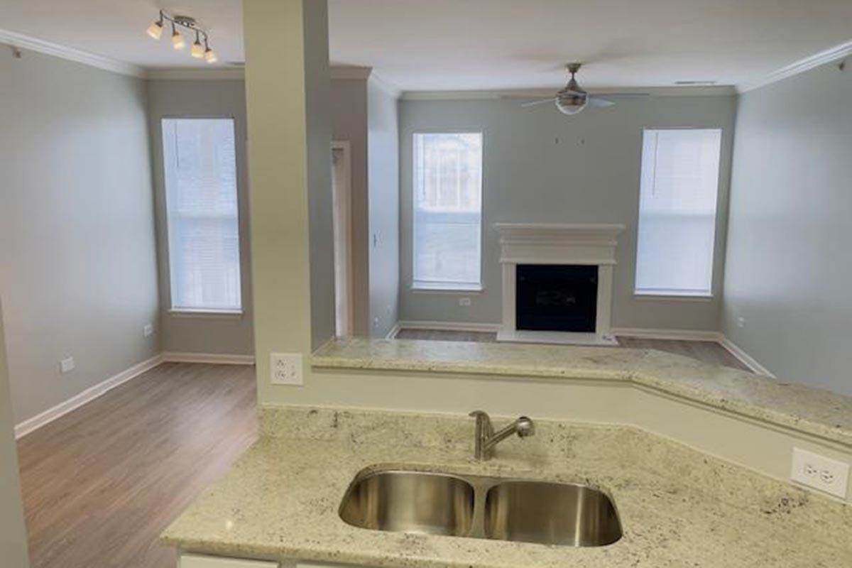 A spacious living area with light gray walls and hardwood flooring, featuring large windows that let in natural light. In the foreground, a kitchen area with a granite countertop and a double sink. A decorative fireplace is visible in the background, enhancing the cozy atmosphere.