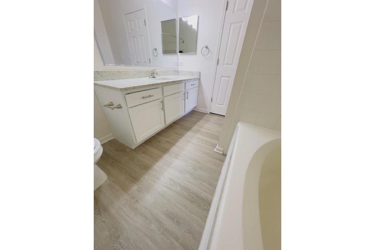 A modern bathroom with a light-colored vanity and countertops. The floor features light wood-style vinyl planks. A large mirror is mounted above the sink, and there is a white bathtub in the corner. The room is bright and clean, with a door leading to another area.
