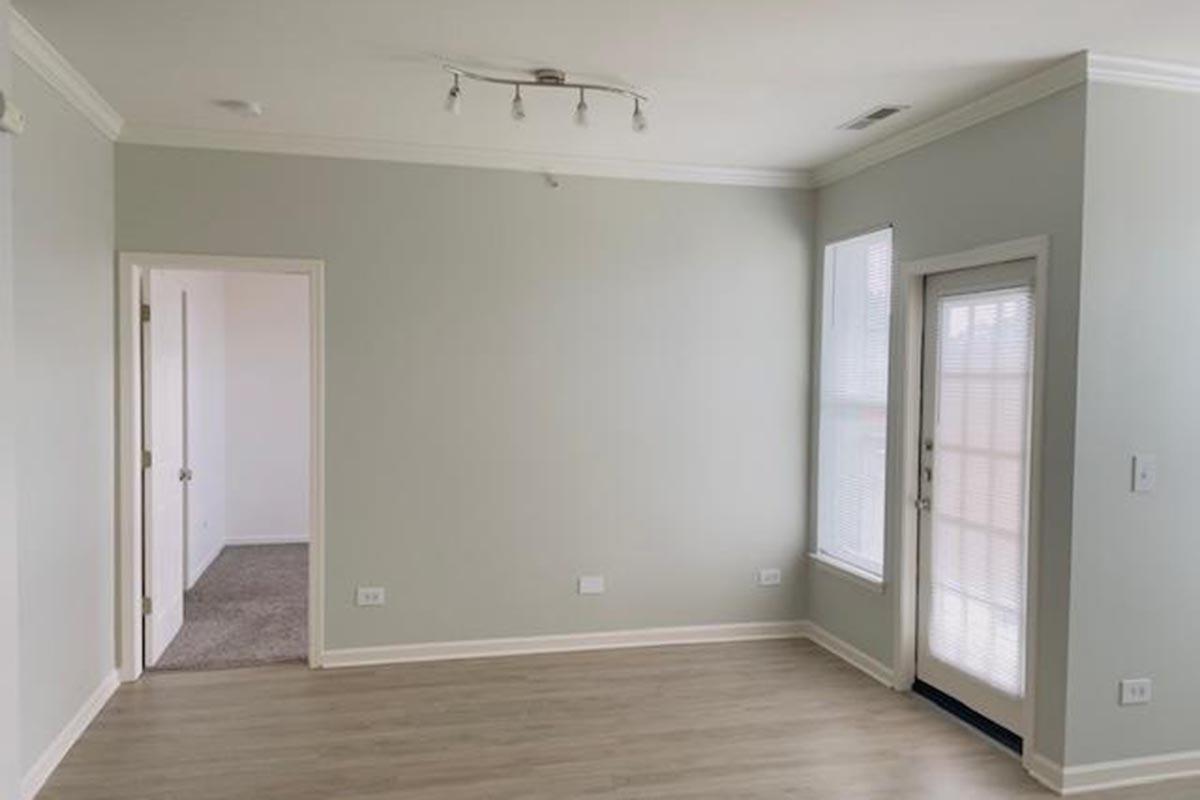 A light-colored room with laminate flooring, featuring a ceiling light fixture and a large window with blinds. A door leads to a small patio, and there is an open doorway to another room. The walls are painted a soft gray, creating a spacious and inviting atmosphere.