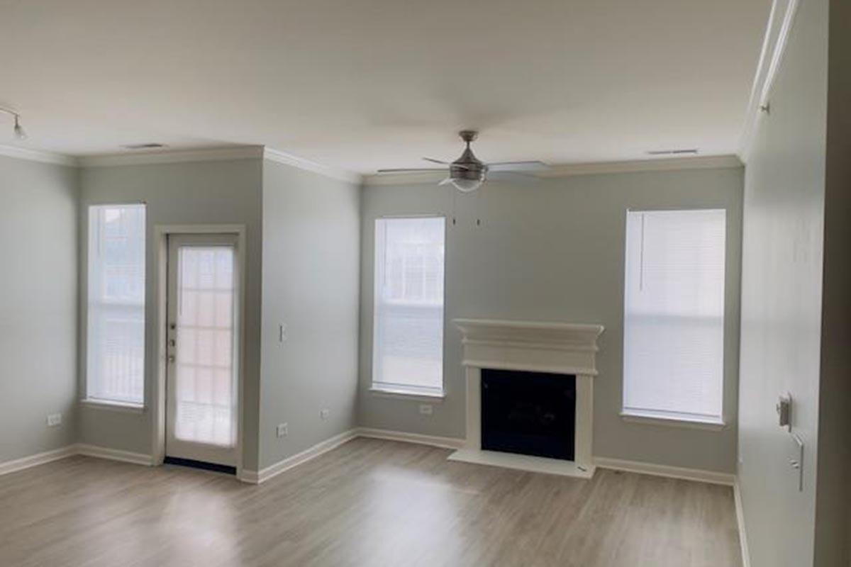 A spacious, light-filled living room featuring pale green walls, two large windows with blinds, and a white fireplace. The room has a ceiling fan and light-colored wooden flooring, creating a modern and inviting atmosphere. A door leads outside, adding to the bright and airy feel.