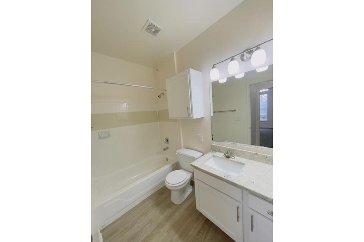 Bright and clean bathroom featuring a bathtub with a shower, white fixtures, a modern vanity with a sink, and a mirror with three light fixtures. Light-colored walls and wood-like flooring create a spacious and inviting atmosphere. Natural light is visible from a window.