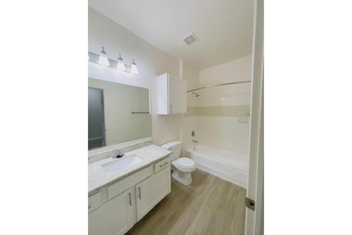 A modern bathroom featuring a white sink, mirror above a countertop, a toilet, and a bath-shower combination. The walls are painted in soft tones, and the floor has light wood-like tiles. Three light fixtures are mounted above the mirror, providing bright illumination.