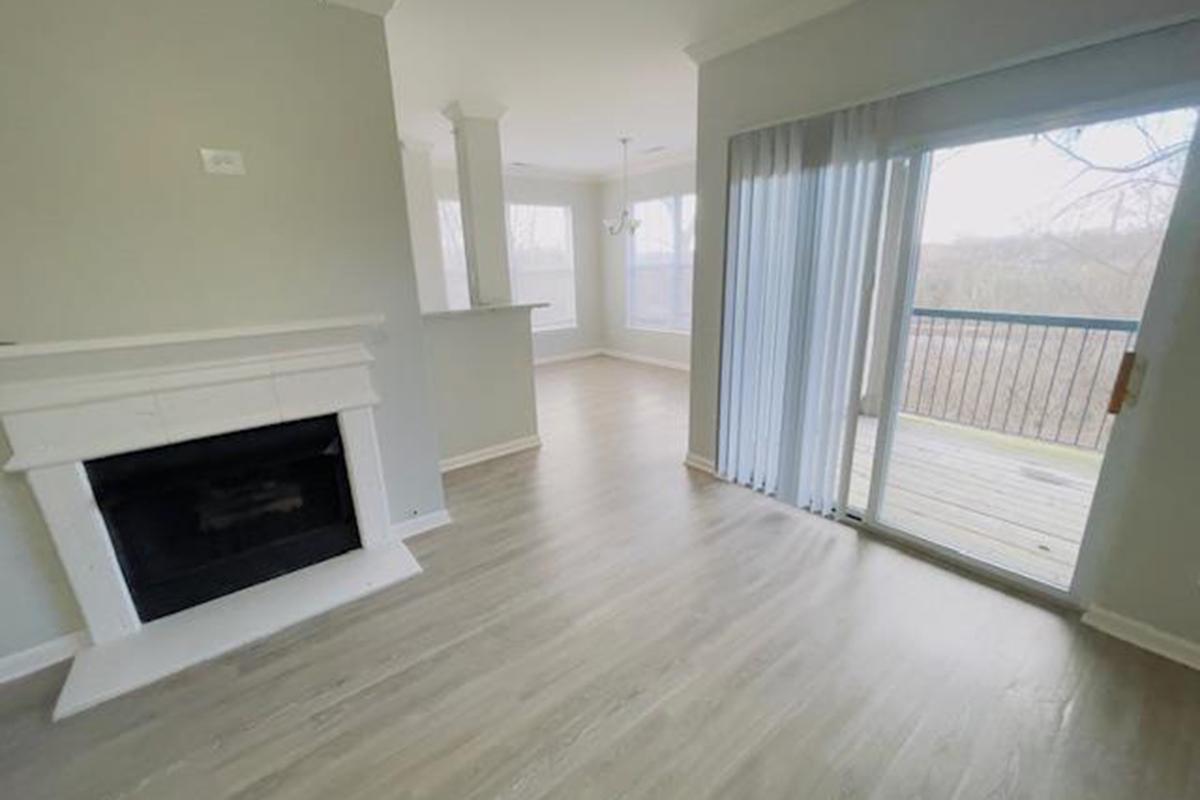 A bright living room featuring light-colored walls and hardwood flooring. There's a fireplace on one side, and large windows with sliding glass doors that lead to a wooden balcony overlooking a natural landscape. The space is open and airy, with natural light flooding in from the windows.
