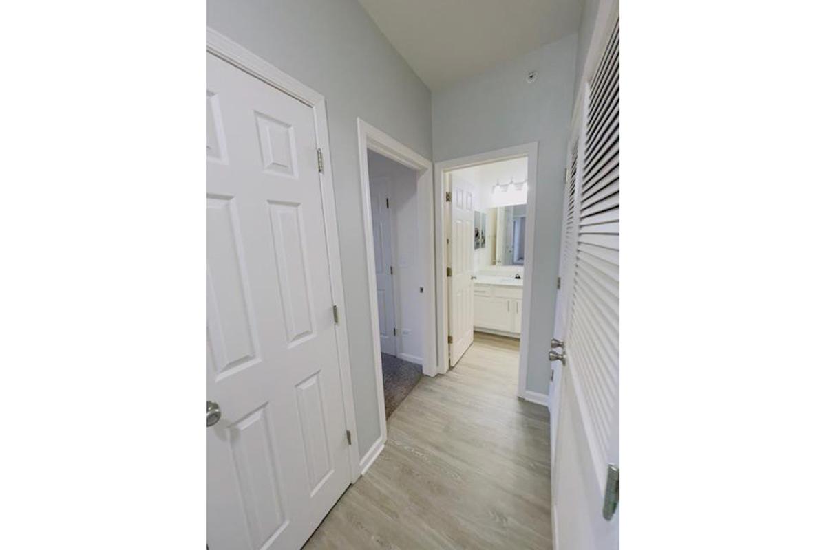Narrow hallway view with white doors leading to other rooms. Light-colored walls and flooring, with a bathroom visible at the end. Soft lighting enhances the clean, modern aesthetic of the space.