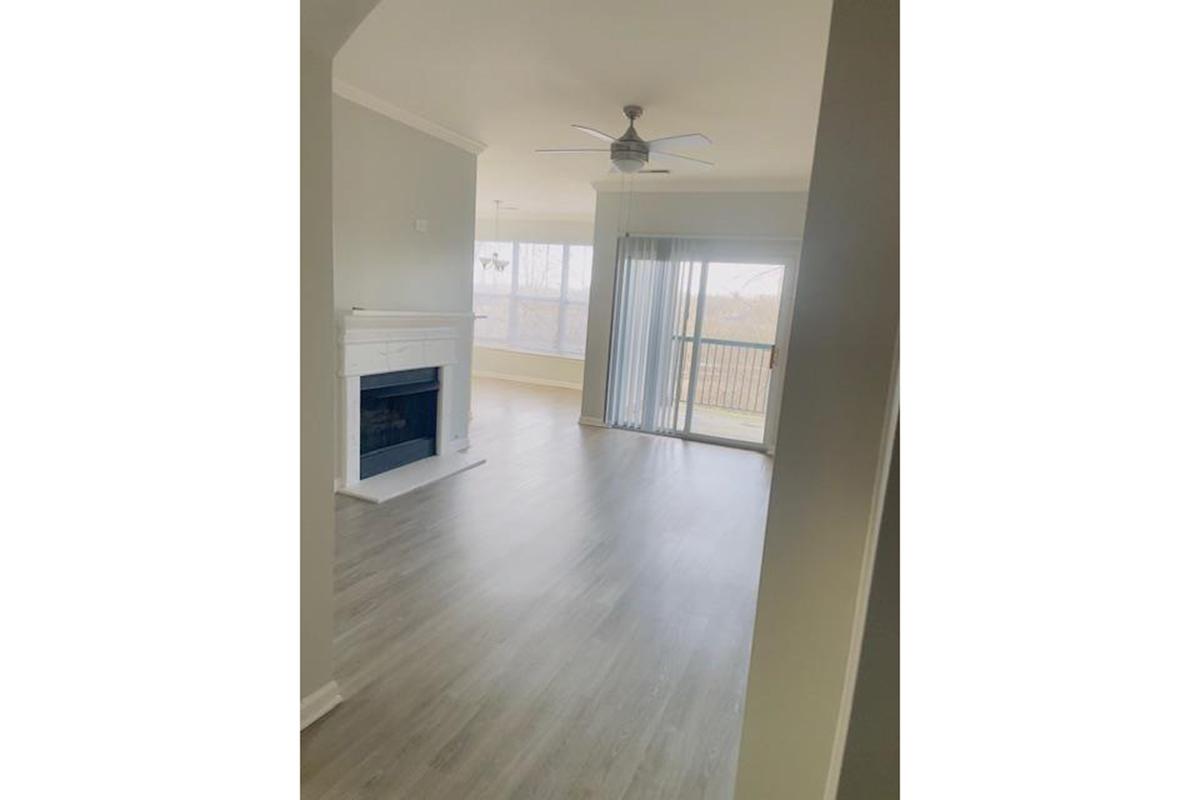 A well-lit, spacious living room featuring a modern ceiling fan, a white fireplace, and large windows with sliding doors leading to a balcony. The flooring is light-colored wood, enhancing the open feel of the space.