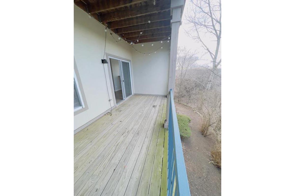 Empty wooden deck with a railing, string lights hanging from the ceiling, and sliding glass doors. The view includes a sloped area with sparse vegetation and trees in the background, set in a cloudy atmosphere.