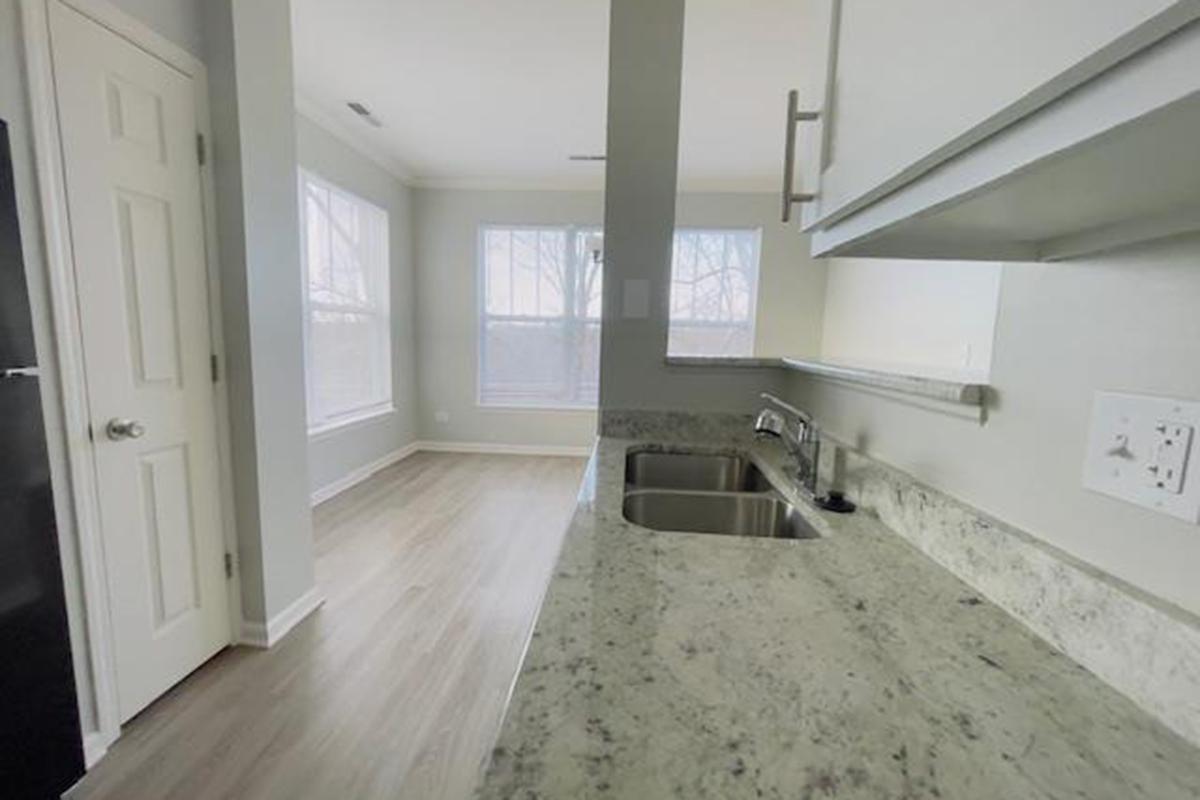 Modern kitchen featuring a granite countertop with double sink, white cabinetry, and a clean design. The background shows a spacious living area with large windows, allowing natural light to fill the room. The flooring is light wood, enhancing the overall bright and airy atmosphere.