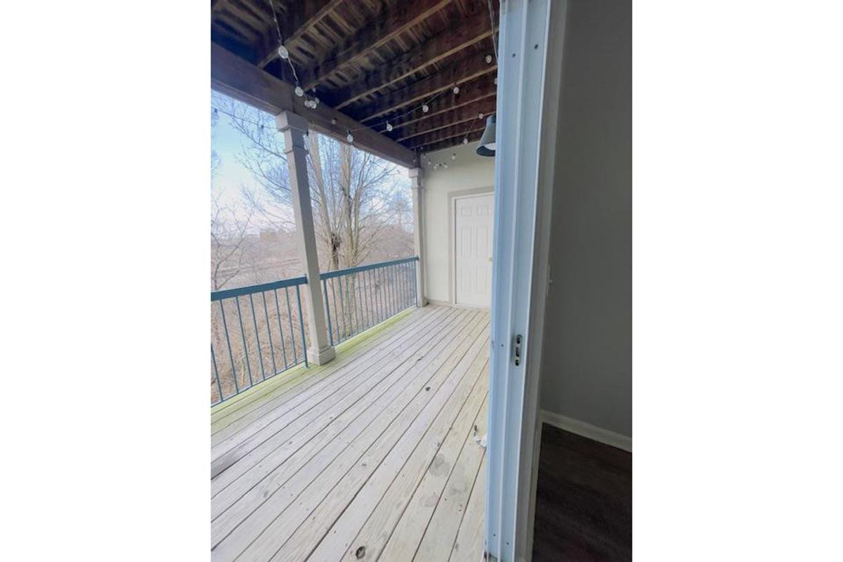 A spacious wooden balcony with a railing, overlooking trees in the background. The entrance door is partially open, inviting light and outdoor views. The floor is made of planks, and the walls are painted in neutral colors, creating a calm and airy atmosphere.