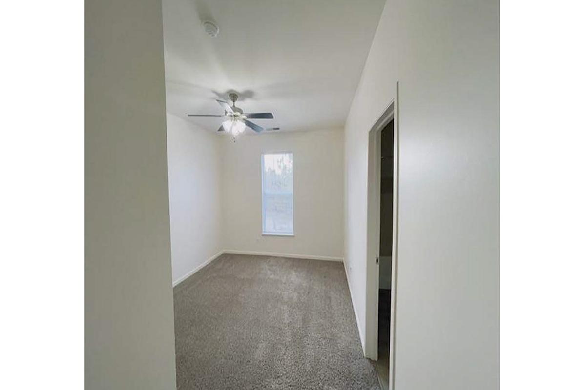 A well-lit, empty room featuring light-colored walls and a ceiling fan. The floor is covered with gray carpet, and there is a window allowing natural light to enter. A doorway leads to another area, suggesting potential for use as a bedroom or office space.