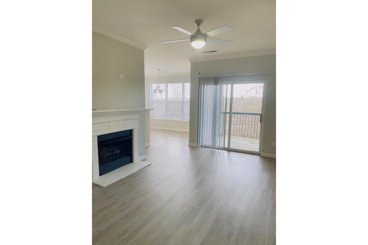 A bright, modern living room featuring a light-colored wall and laminate flooring. There is a ceiling fan, a large window with sliding glass doors leading to a balcony, and a white fireplace. The space is uncluttered and inviting, showcasing natural light.