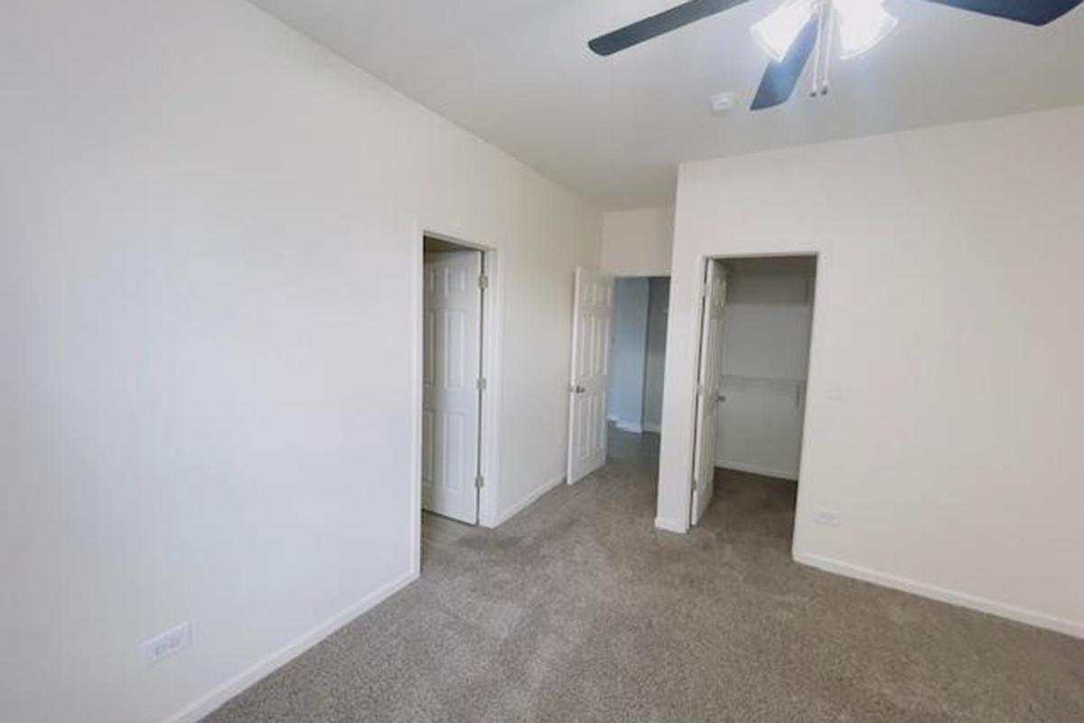 A bright, empty room featuring light-colored walls and a carpeted floor. There are two doors: one leading to a closet and the other likely to another area. A ceiling fan is visible, adding to the spacious feel of the room. The overall ambiance is clean and simple, suitable for a variety of uses.