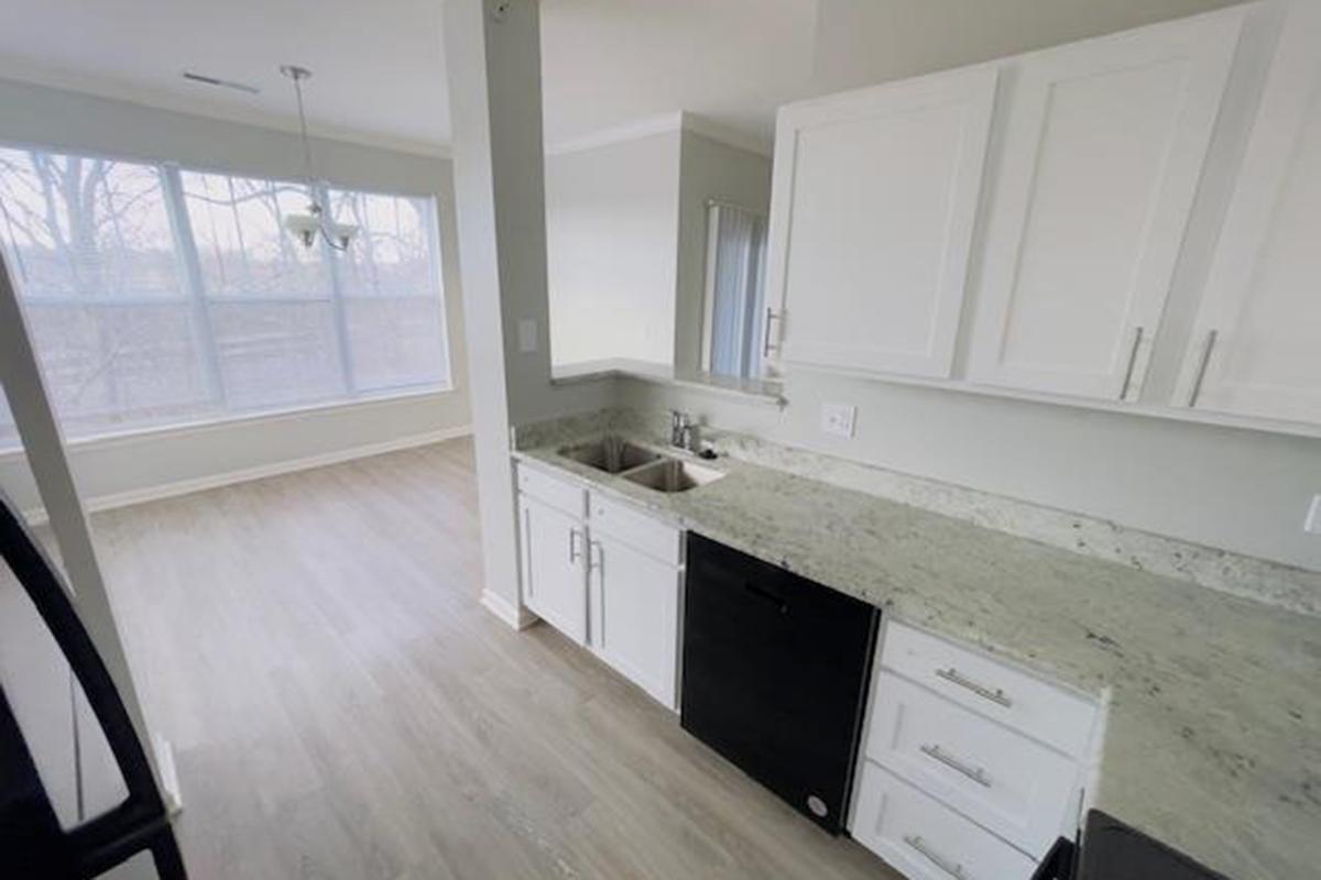 Modern kitchen with white cabinets and gray granite countertops. Features a sink and a black dishwasher. To the right, there is a spacious dining area with large windows allowing natural light in. The flooring is light-colored wood, creating a bright and airy atmosphere.