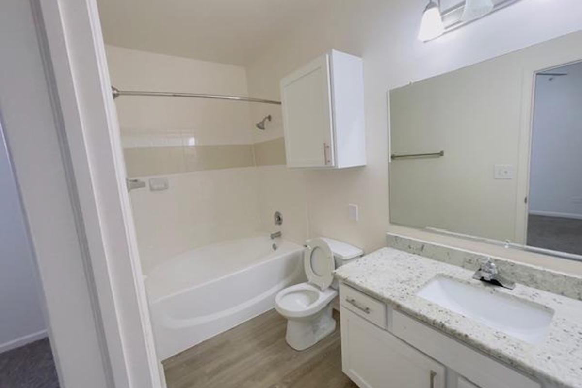 A clean, modern bathroom featuring a white bathtub, toilet, and sink with a granite countertop. The walls are light-colored with a shower rod above the tub. A large mirror and cabinet above the sink provide additional storage. The flooring is light wood-like laminate, contributing to the contemporary feel of the space.