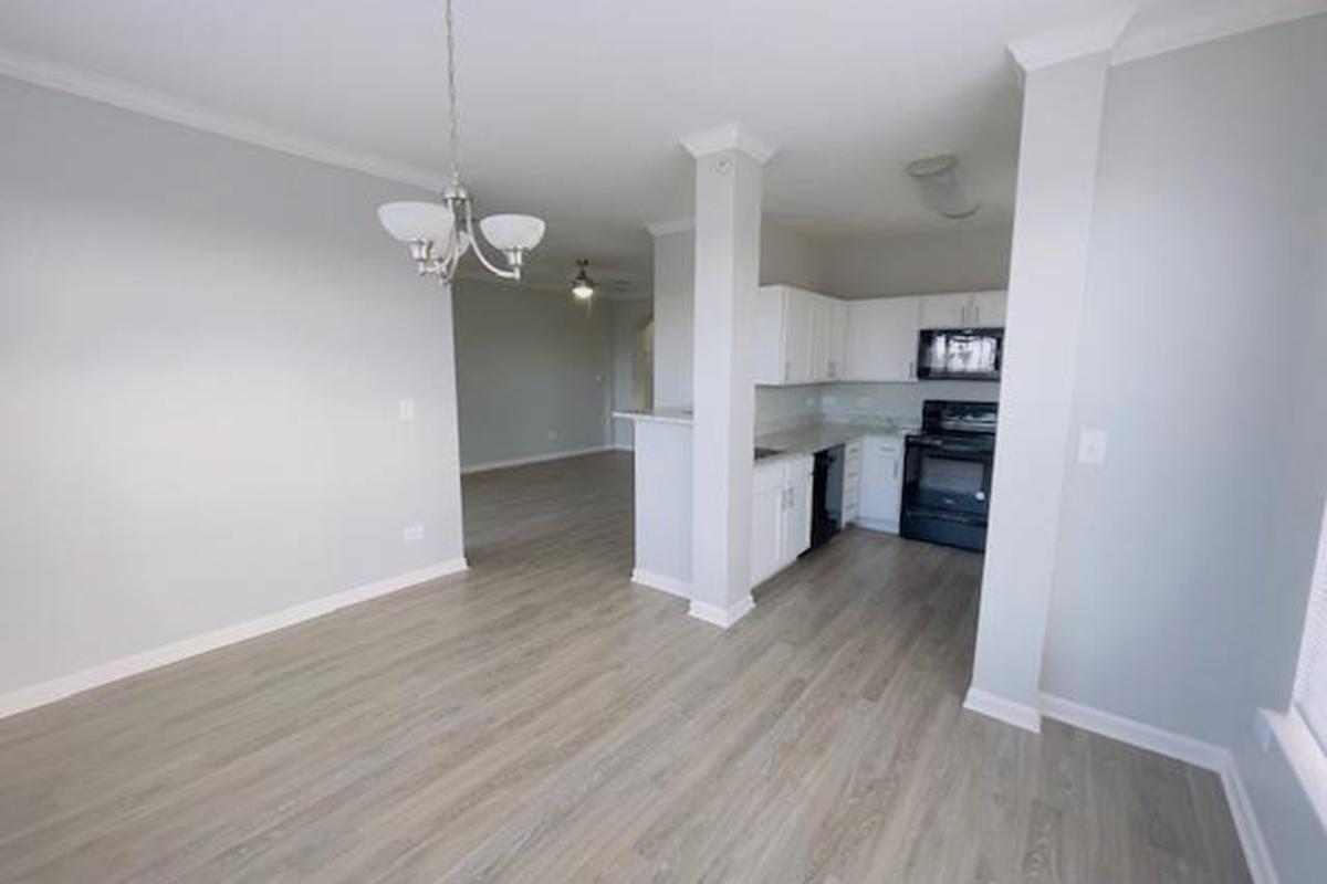 A modern, open-concept kitchen and dining area featuring light gray walls, a chandelier, and wooden flooring. The kitchen includes white cabinets, a black oven, and countertop space. Natural light illuminates the space, creating a spacious, airy feel.