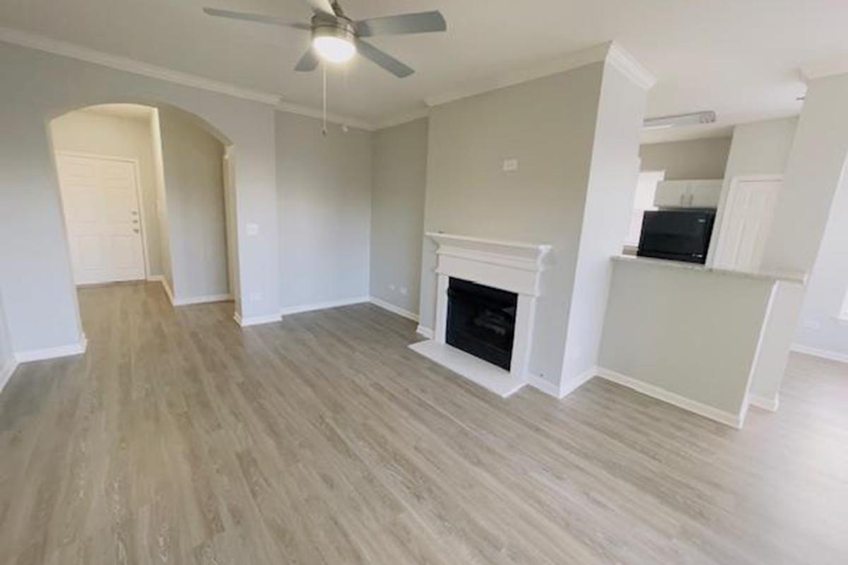 Interior of a modern living room featuring light gray walls, a ceiling fan, and a fireplace. The room has laminate flooring and an open layout leading to a kitchen area. A doorway in the back suggests access to another room or entryway, with a small black microwave visible on the counter.