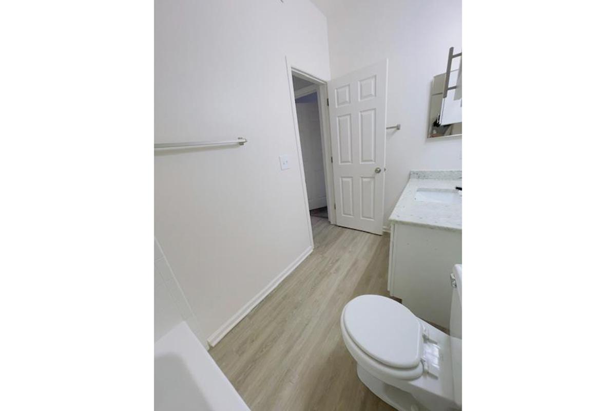 A clean, modern bathroom featuring a toilet, a sink with a countertop, and a light-colored wooden floor. The walls are painted in a neutral tone, and a door leads to another room in the background. Soft natural light fills the space, creating an inviting atmosphere.