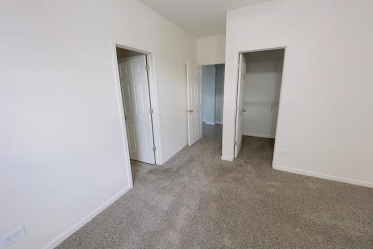 A light-colored room with beige carpet, featuring two open doors on the left leading to other areas. One door opens into a small closet, while the other leads to an adjoining space. The walls are bare, creating a clean and minimalistic look. Natural light may be present, enhancing the room's spacious feel.