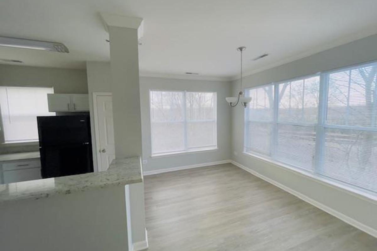 Bright and airy living space featuring large windows with blinds, a light-colored wall, and wood-style flooring. The kitchen area includes dark appliances and a countertop with a granite finish. Sunlight streams in, creating a warm and inviting atmosphere.