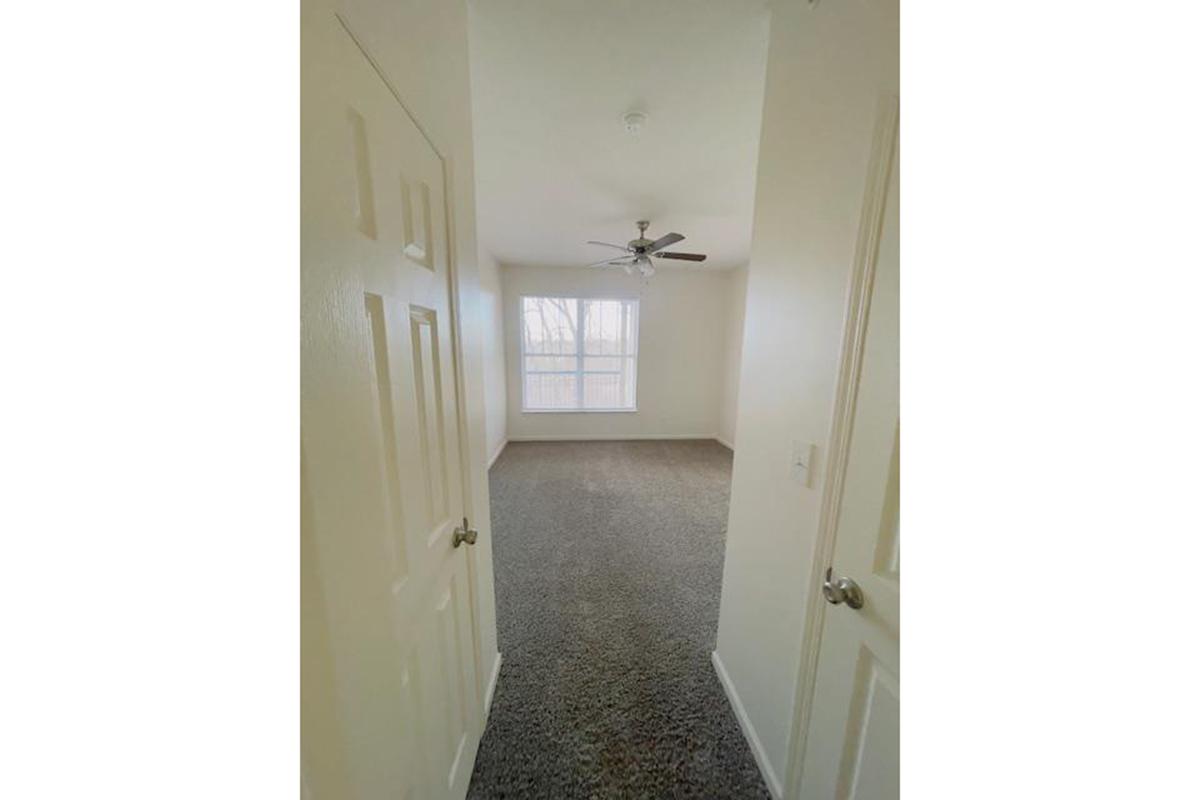 A well-lit room featuring light-colored walls and carpet. It has a ceiling fan and a large window that provides natural light. The image shows a view from the doorway, with two closed doors visible on the left side, leading into the room.
