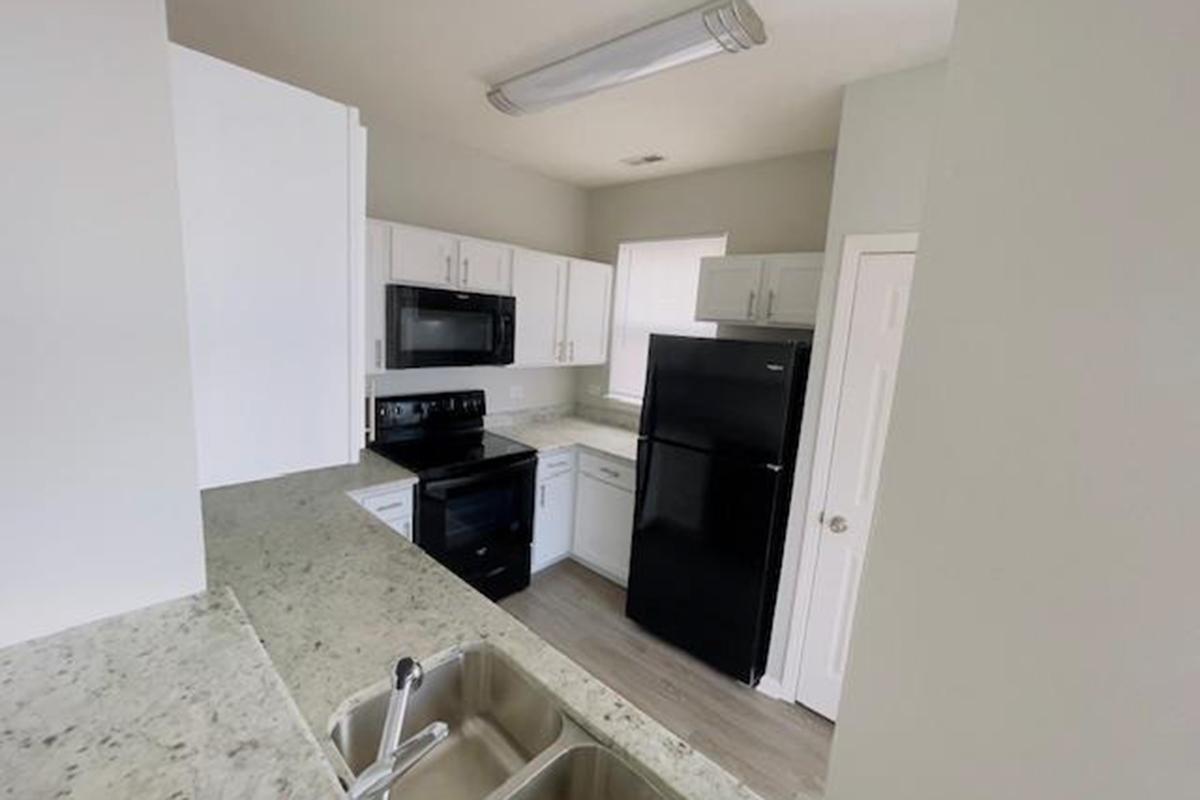 A modern kitchen featuring white cabinets, a black refrigerator, and a microwave. The countertops are light-colored with a speckled design. There is a sink with two basins and a window providing natural light. The flooring is light-colored, enhancing the bright, clean aesthetic of the space.