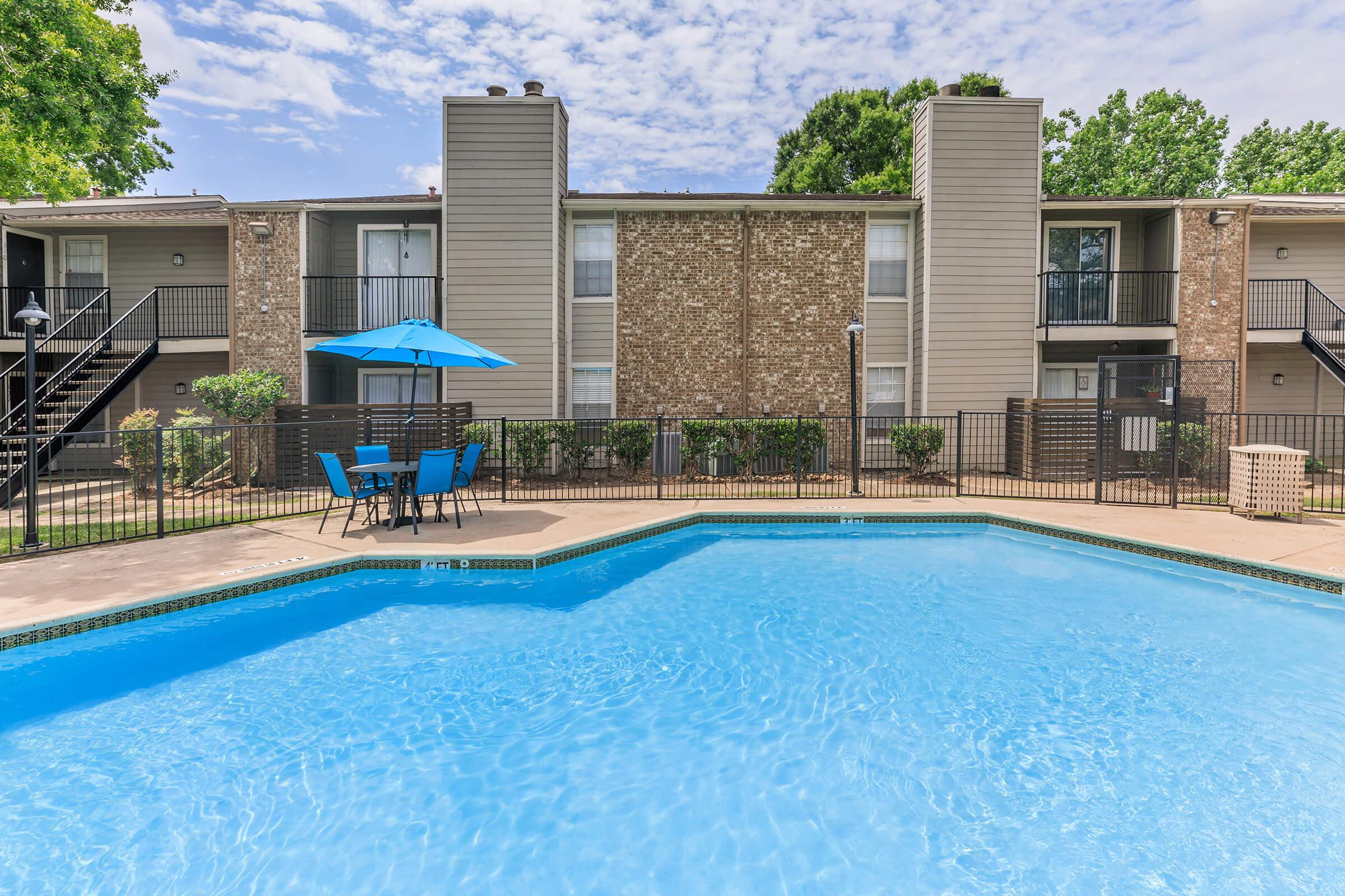 A bright, inviting swimming pool surrounded by a fenced area, with blue lounge chairs and a patio table under a blue umbrella. In the background, there's a two-story apartment building with balconies and greenery. The sky is partly cloudy, creating a sunny atmosphere.