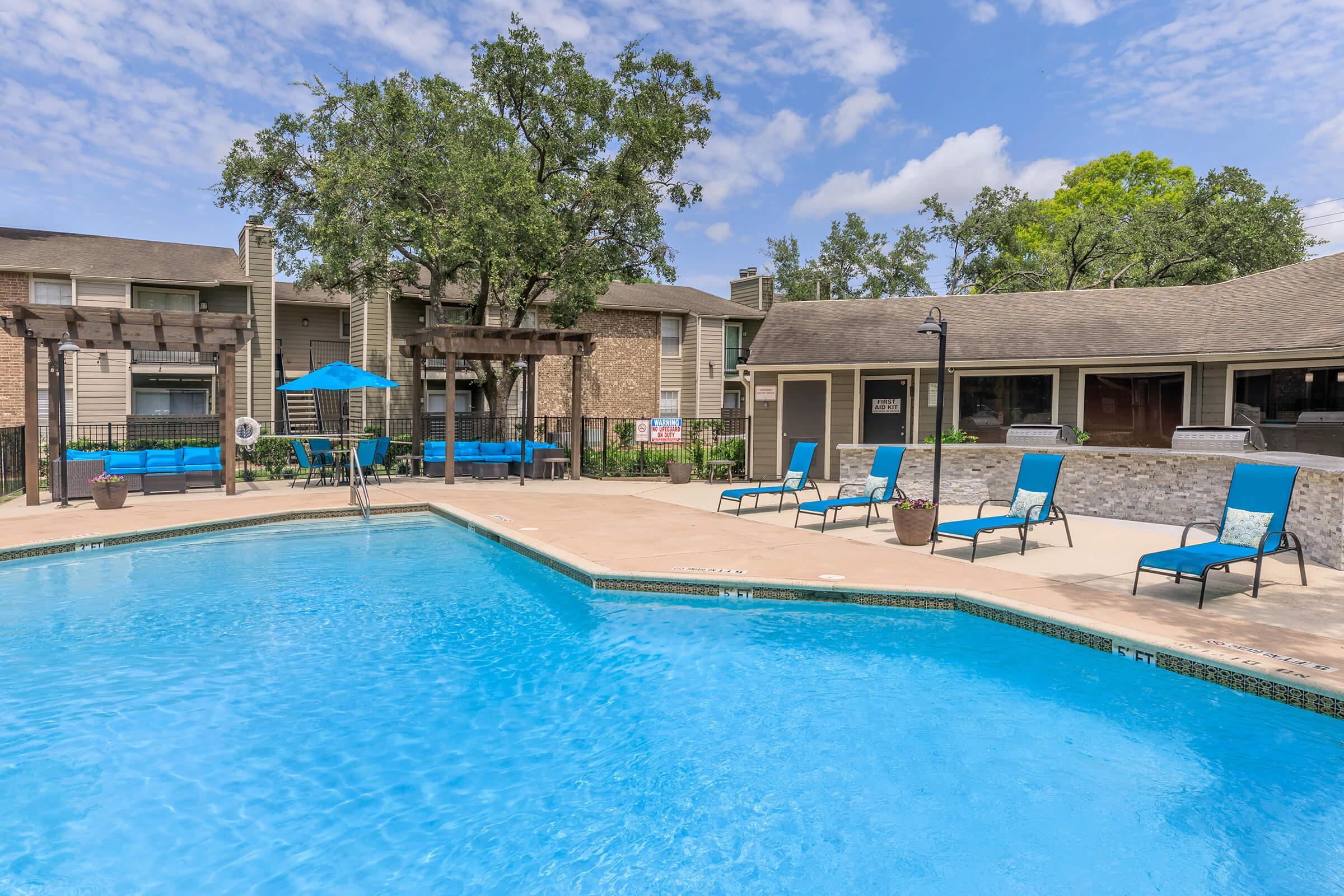 A clear blue swimming pool surrounded by lounge chairs and umbrellas, set in a residential area with trees and well-maintained landscaping. The sky is partly cloudy, and there are buildings with patios in the background.