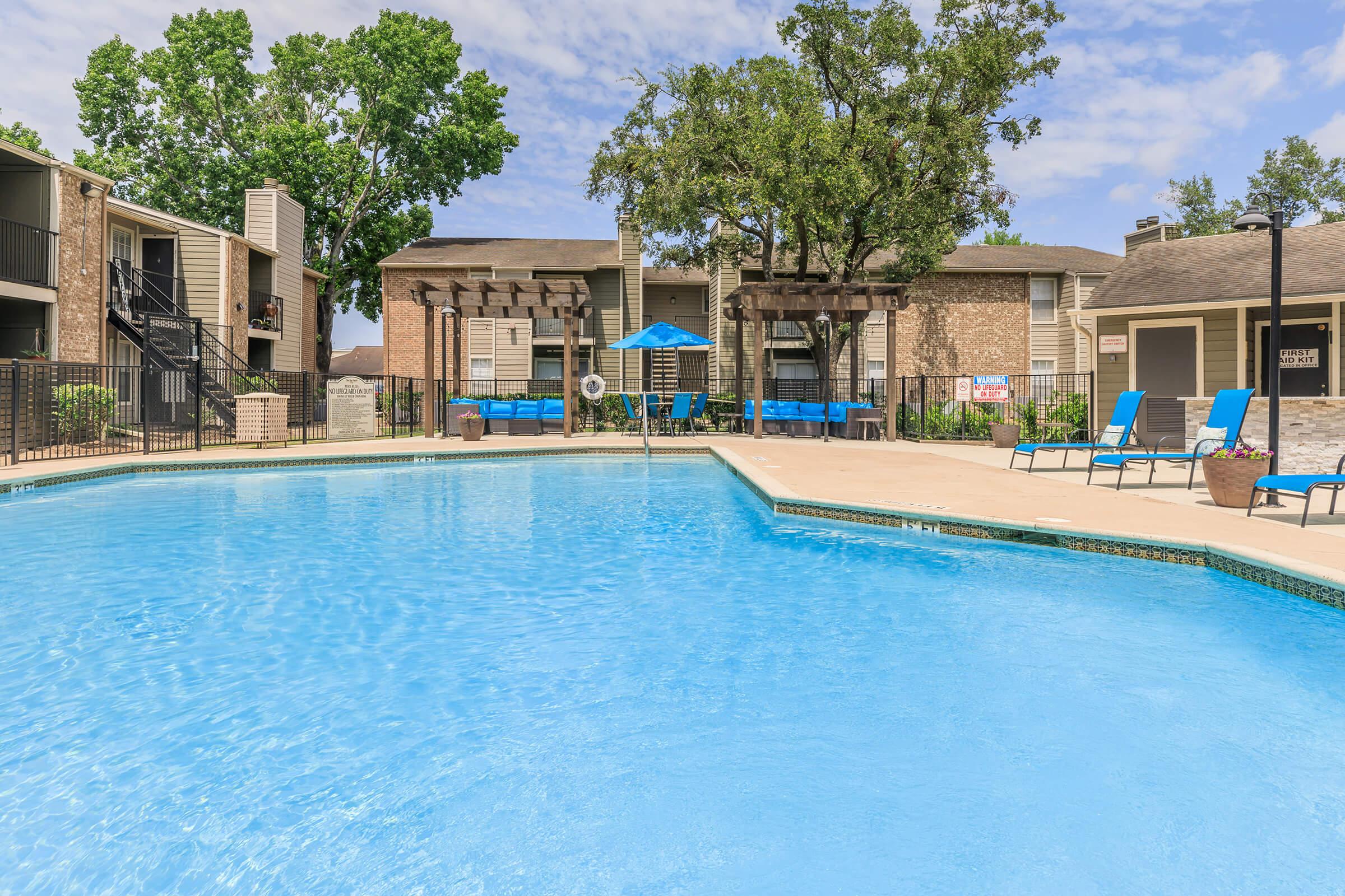 A clear blue swimming pool surrounded by lounge chairs and shaded areas. Apartment buildings with balconies are visible in the background, complemented by trees and a blue sky with scattered clouds. The scene conveys a relaxing outdoor environment.