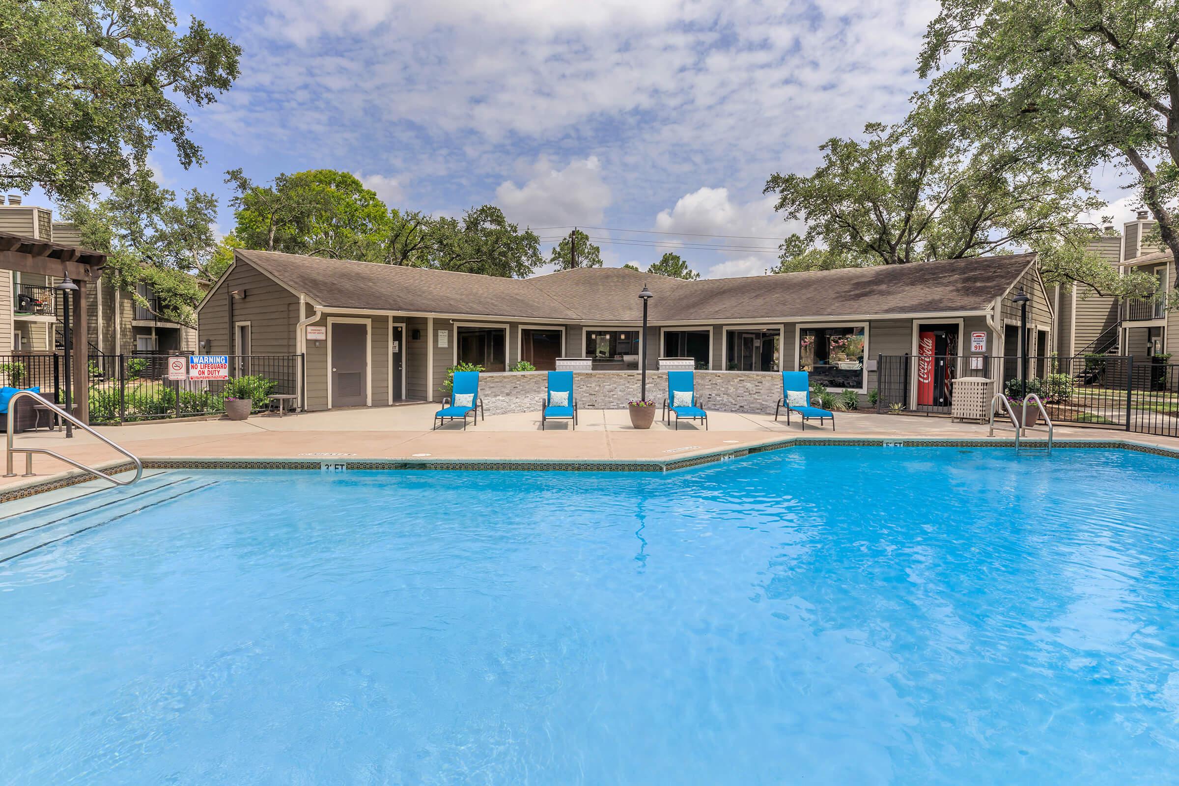 A clear blue swimming pool in the foreground, bordered by lounge chairs. In the background, a building with large windows and a sloped roof, surrounded by trees under a partly cloudy sky. The setting suggests a relaxed outdoor environment ideal for leisure and recreation.