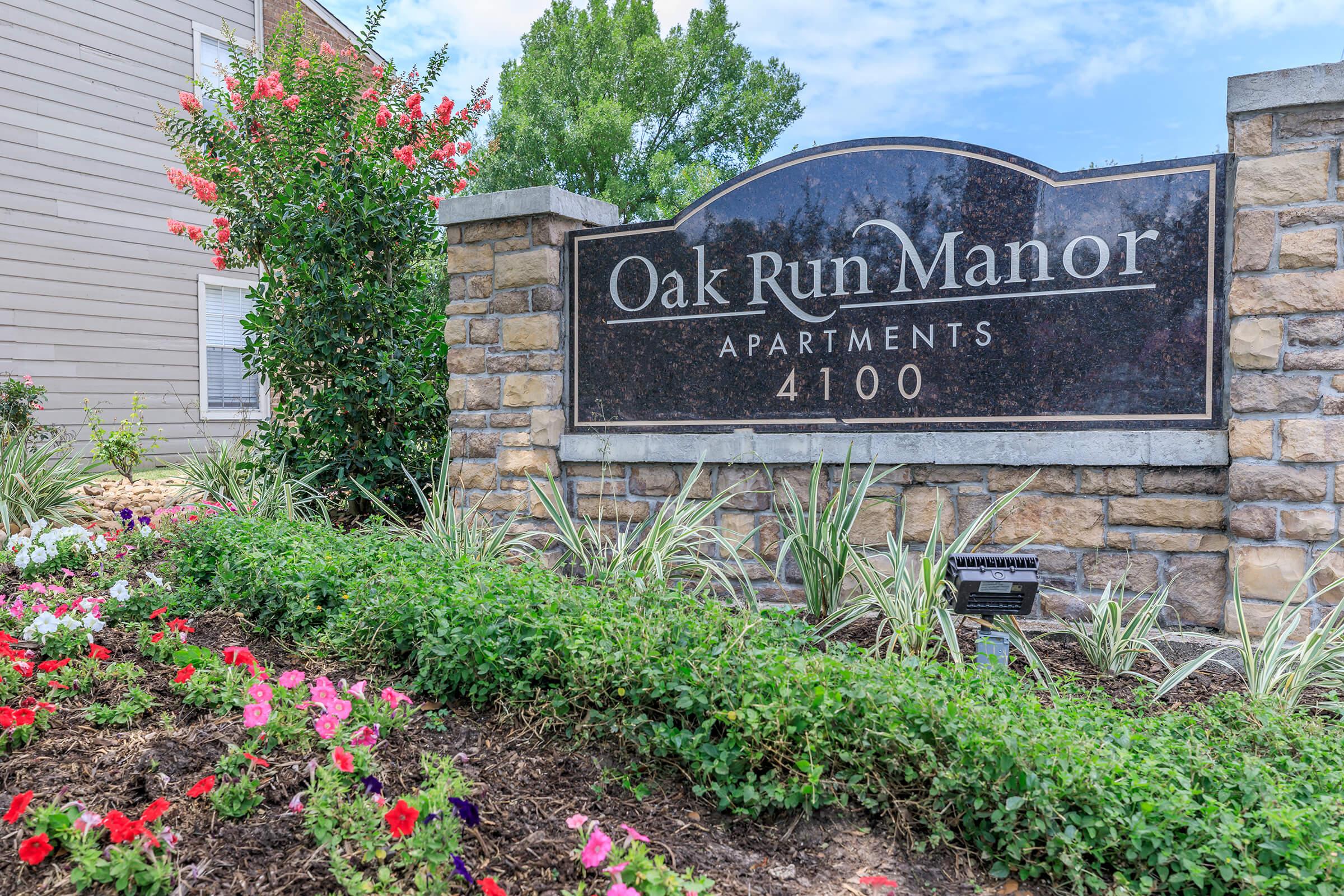 Sign for Oak Run Manor Apartments, featuring stone and dark marble design. Surrounding the sign are colorful flower beds with various plants, adding a vibrant touch to the entrance. The apartment complex number is displayed as 4100. Bright blue sky and greenery are visible in the background.