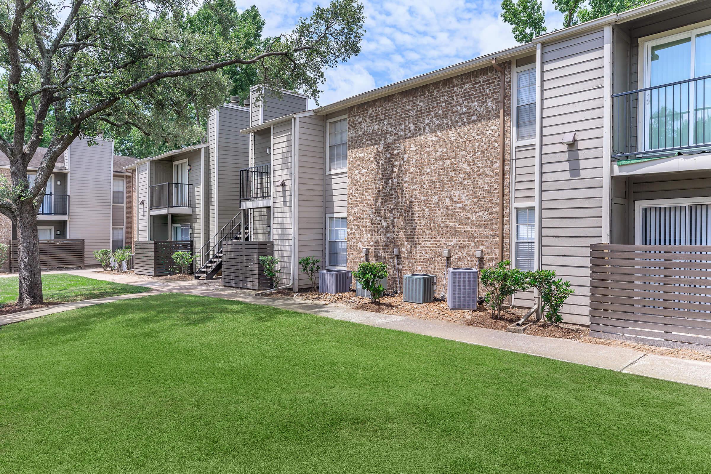 A landscaped area featuring two multi-level apartment buildings. The buildings have wood and brick exteriors with balconies. Lush green grass in the foreground, surrounded by shrubs and small planters. Bright blue sky with scattered clouds overhead.