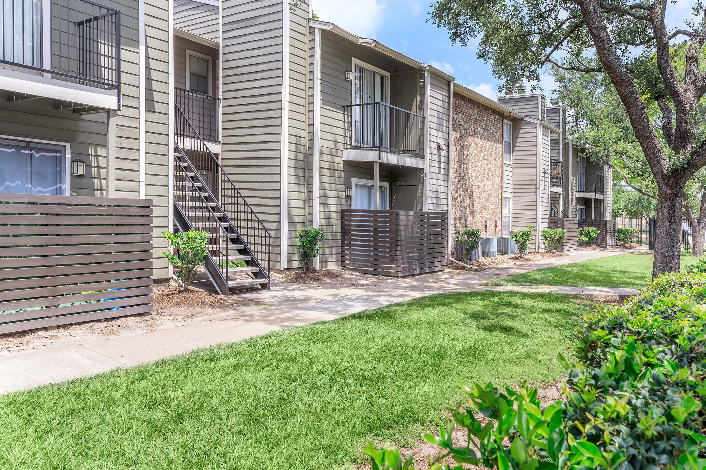 A well-maintained apartment complex features multiple buildings with balconies, surrounded by lush green grass and neatly trimmed hedges. A paved walkway runs alongside the buildings, leading to staircases at the entrances. Trees provide shade, enhancing the inviting atmosphere of the outdoor space.