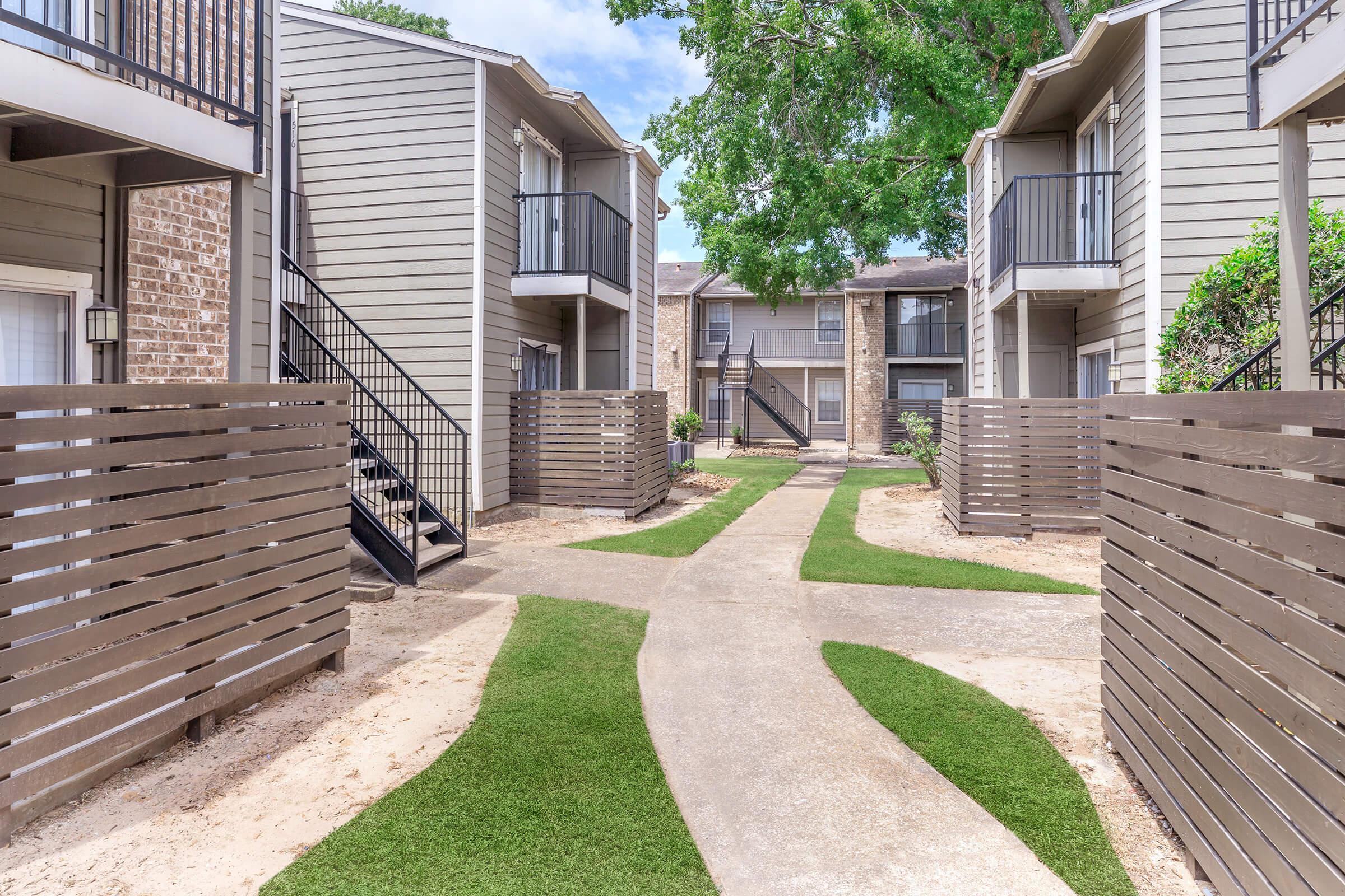 A well-maintained apartment complex courtyard featuring two-story buildings with balconies. The pathway is lined with green grass and bordered by wooden fencing, leading to an inviting, open area surrounded by greenery. The scene is bright and sunny, creating a welcoming atmosphere.